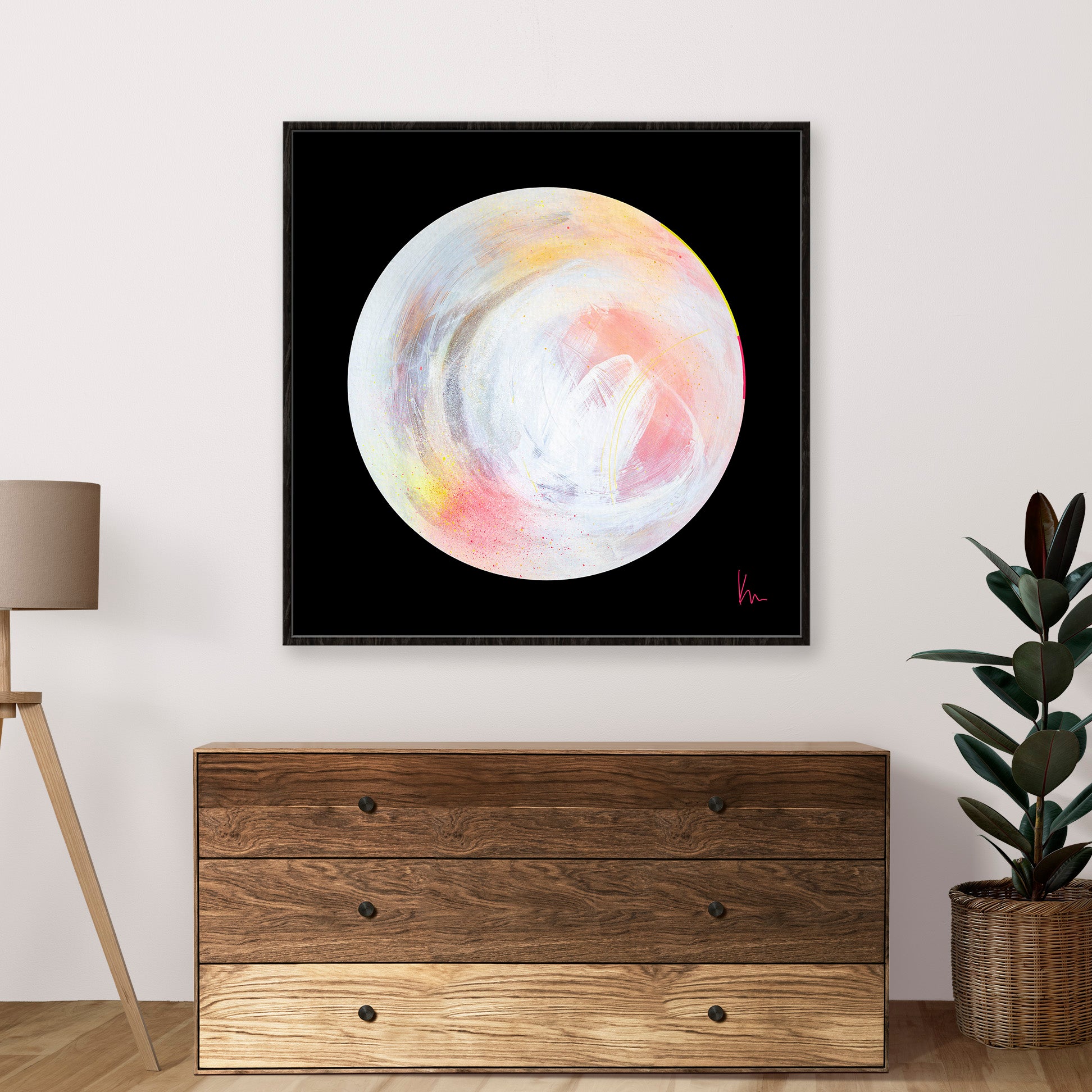 The Time Is Now," a framed abstract circle painting in pastel colors by the artist Kate Mayer, is displayed above a wooden dresser. A lamp and plant are situated on either side of the dresser.