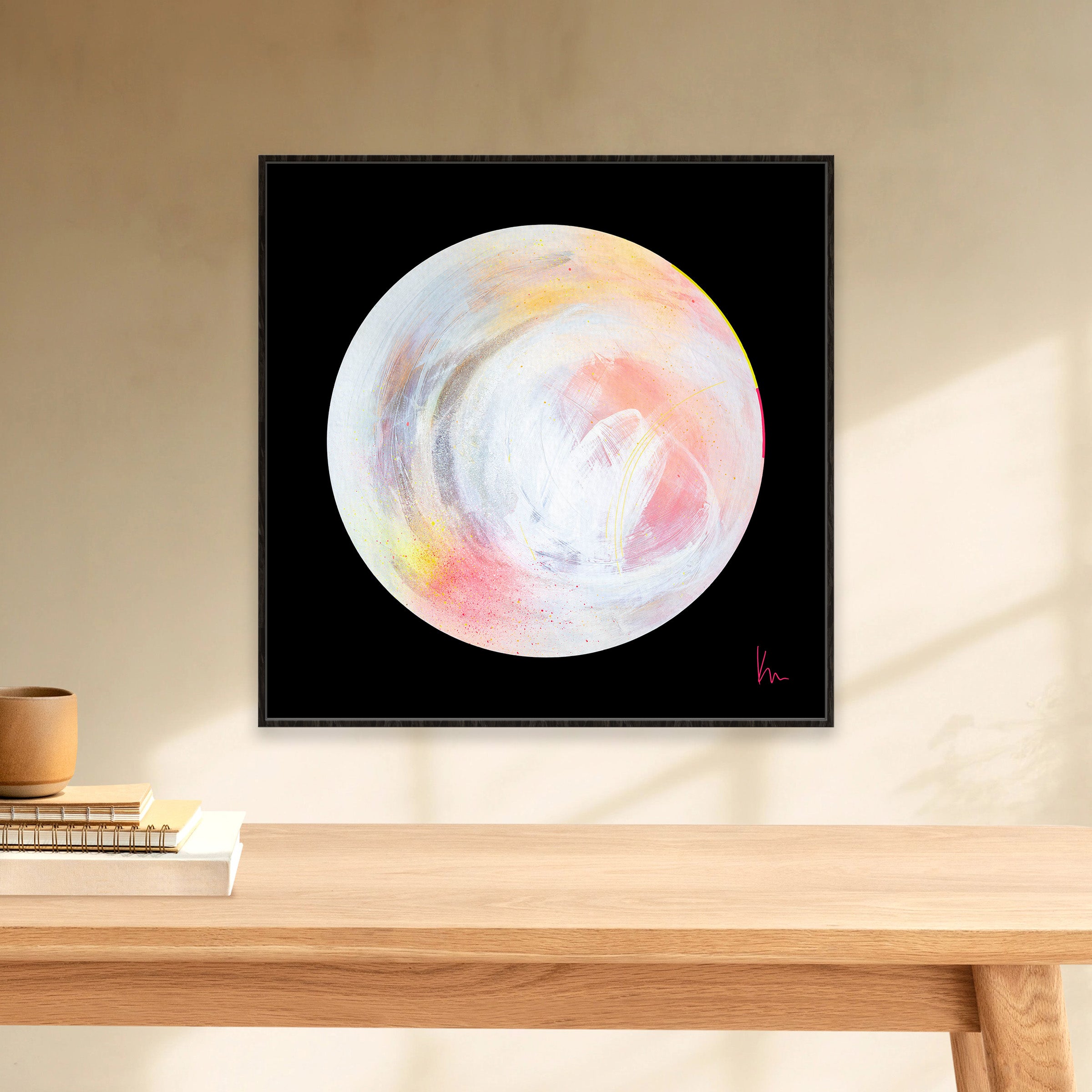 A framed painting by Kate Mayer, titled "The Time Is Now," featuring a white and pastel-colored circle on a black background, is hung on a light-colored wall above a wooden shelf.