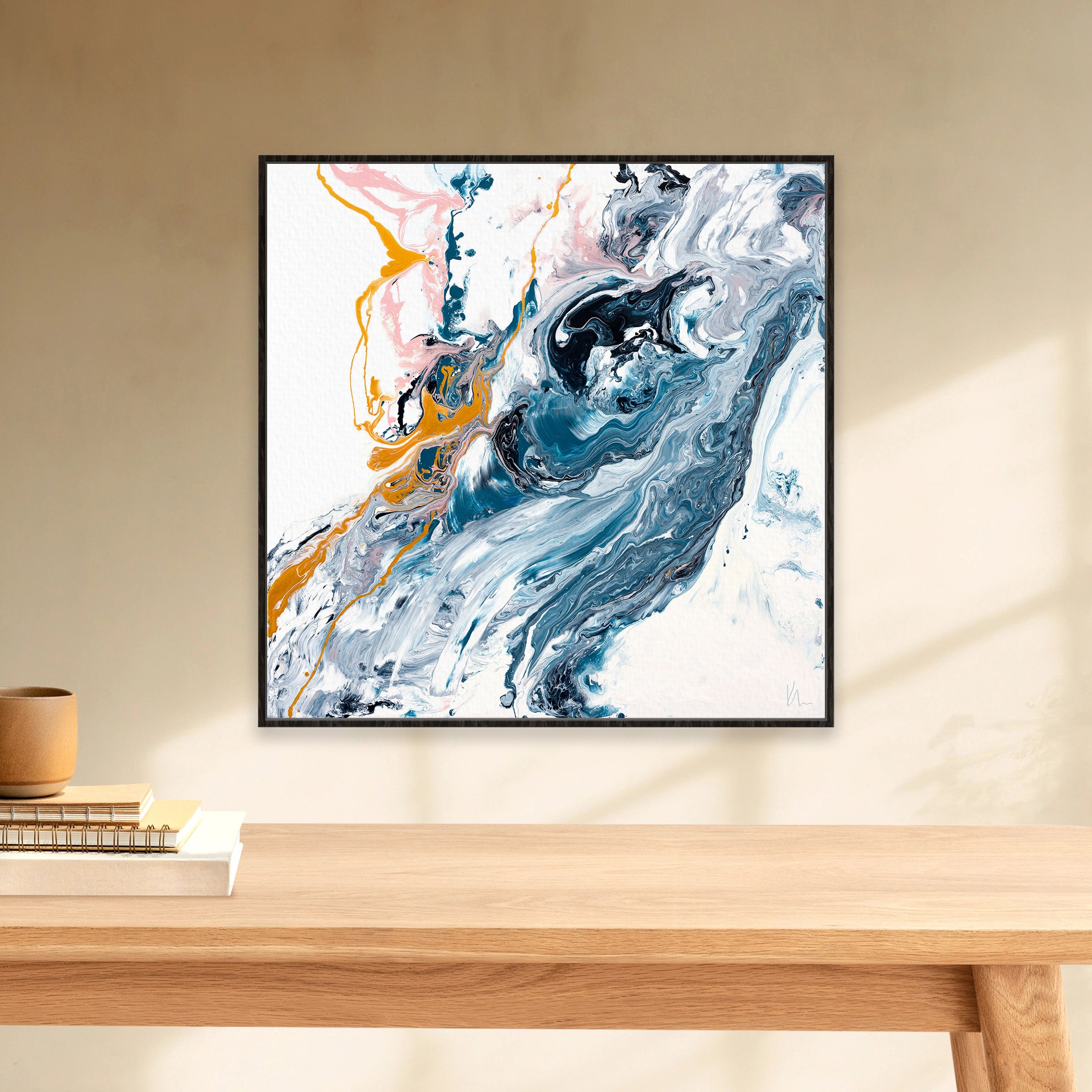 Kate Mayer's "Undercurrent" abstract painting, featuring swirling blue, white, pink, and yellow hues in a sleek black frame, hangs on a beige wall above a wooden table.