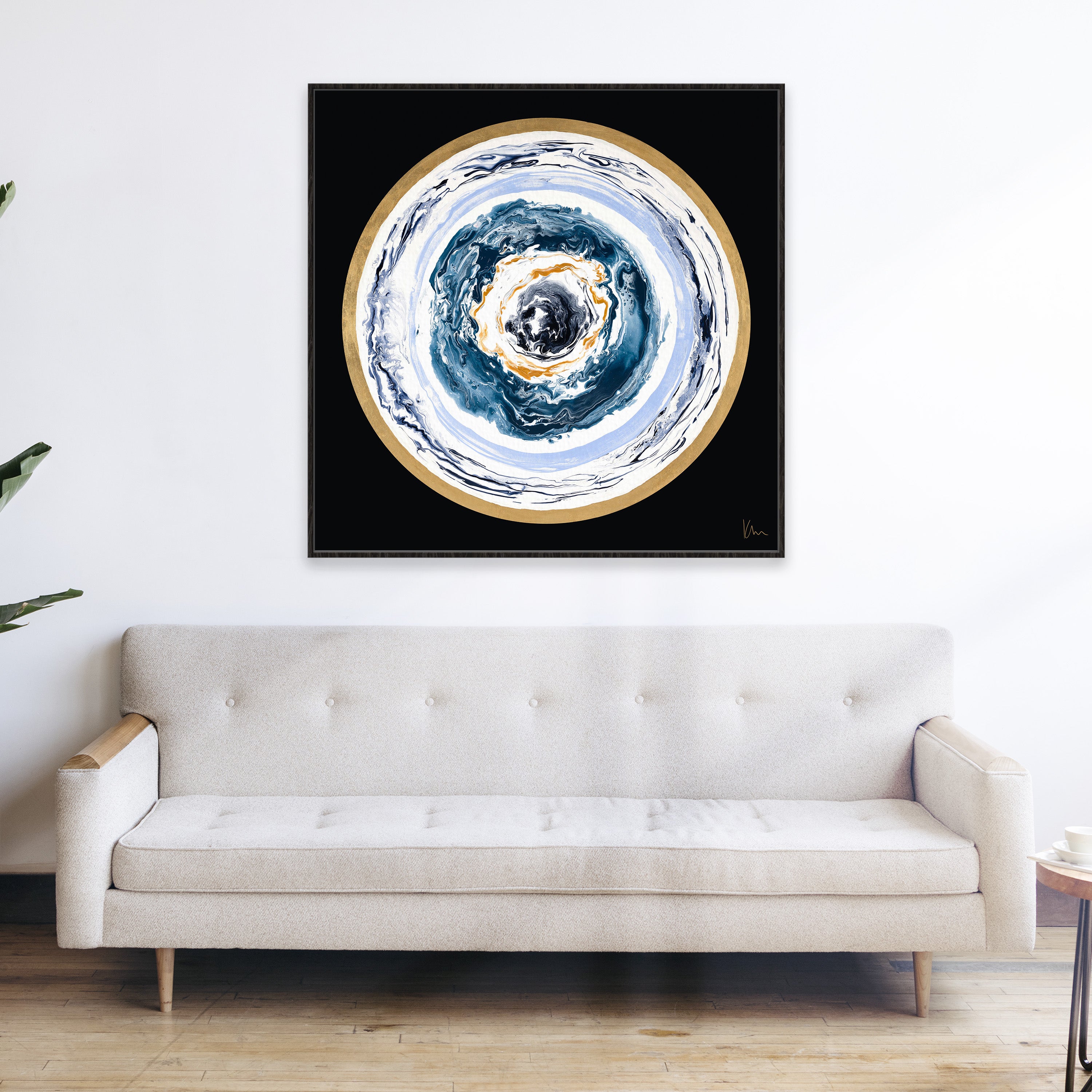 Modern living room with a white sofa and "Universal Love," a circular abstract painting in blue, white, and gold tones by Kate Mayer, hanging on the wall above. This description encapsulates a contemporary yet elegant vibe.