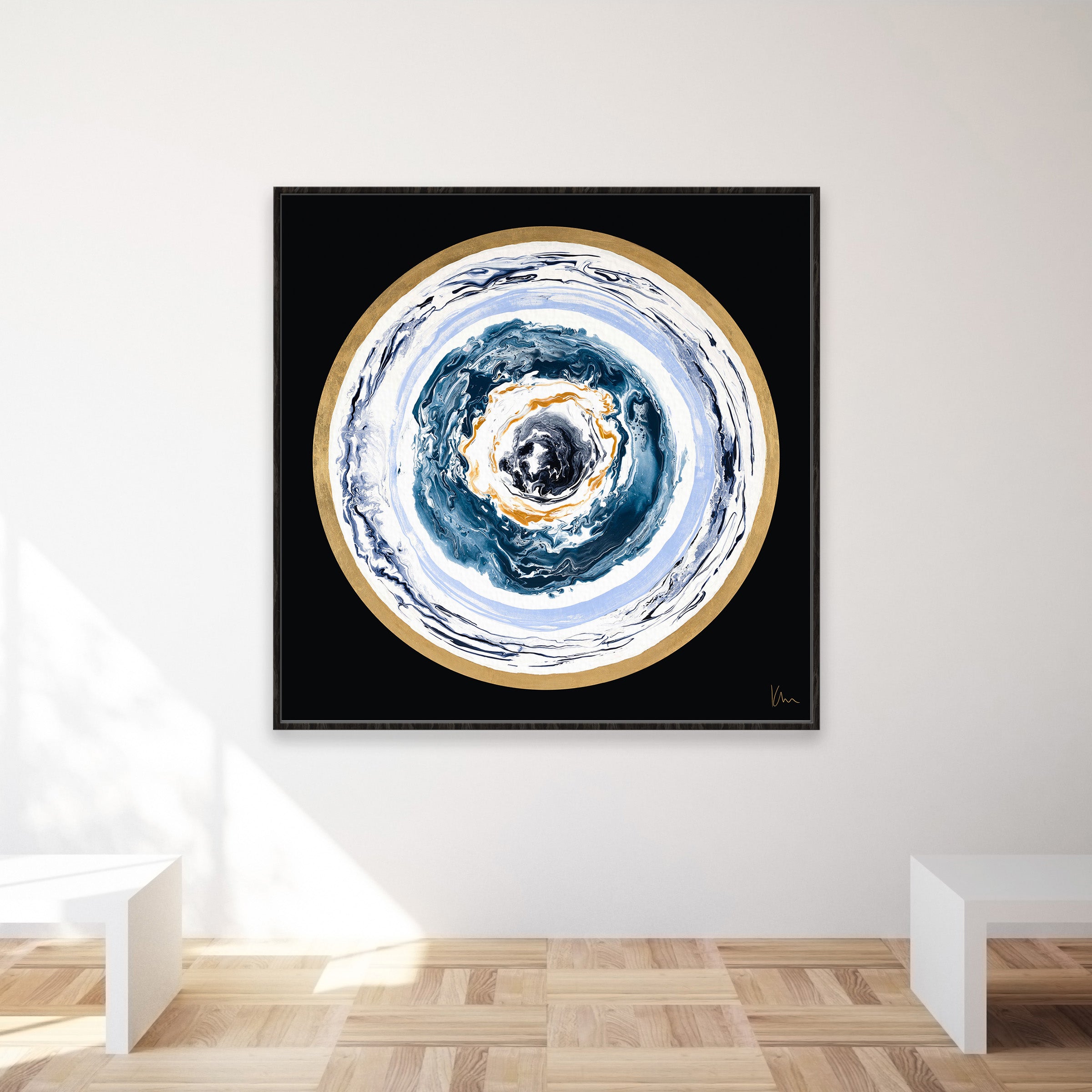 A minimalistic room with wooden flooring features "Universal Love," a square abstract painting by Kate Mayer, showcasing concentric circular patterns in black, white, and gold. This description captures the serene ambiance and artistic flair of the space.