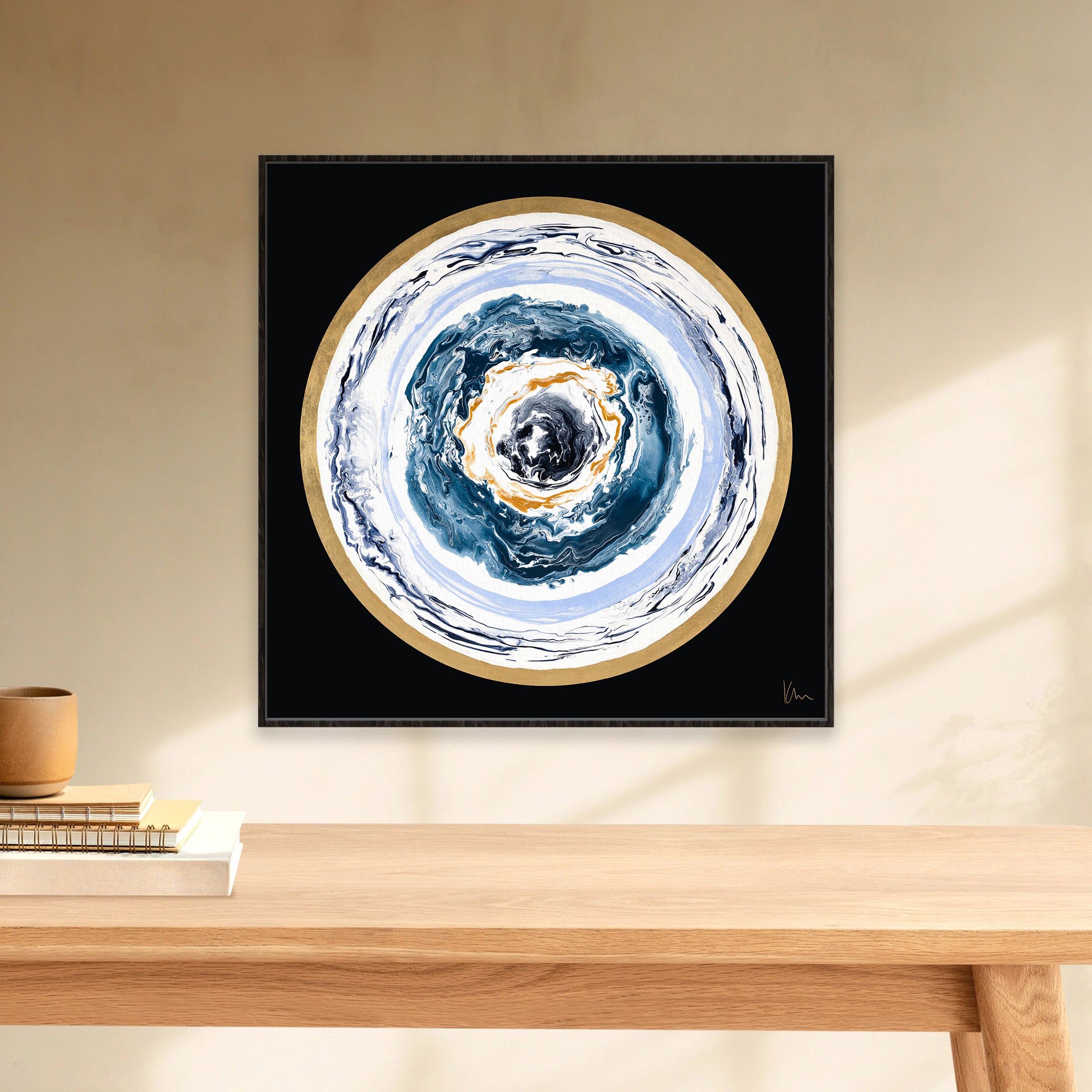 A mesmerizing abstract circular painting titled "Universal Love" by Kate Mayer, featuring blue, white, and gold swirls, is mounted on a beige wall above a wooden table decorated with books and a plant.