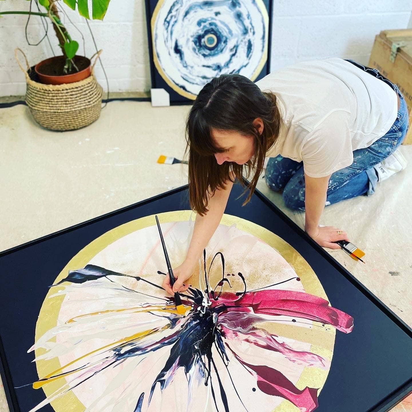 A person kneels to paint an abstract artwork on a black canvas, incorporating acrylic and gold leaf. In the background, an Expansions - Original Painting by Kate Mayer hangs ready-to-hang next to a potted plant, contributing to the serene atmosphere.