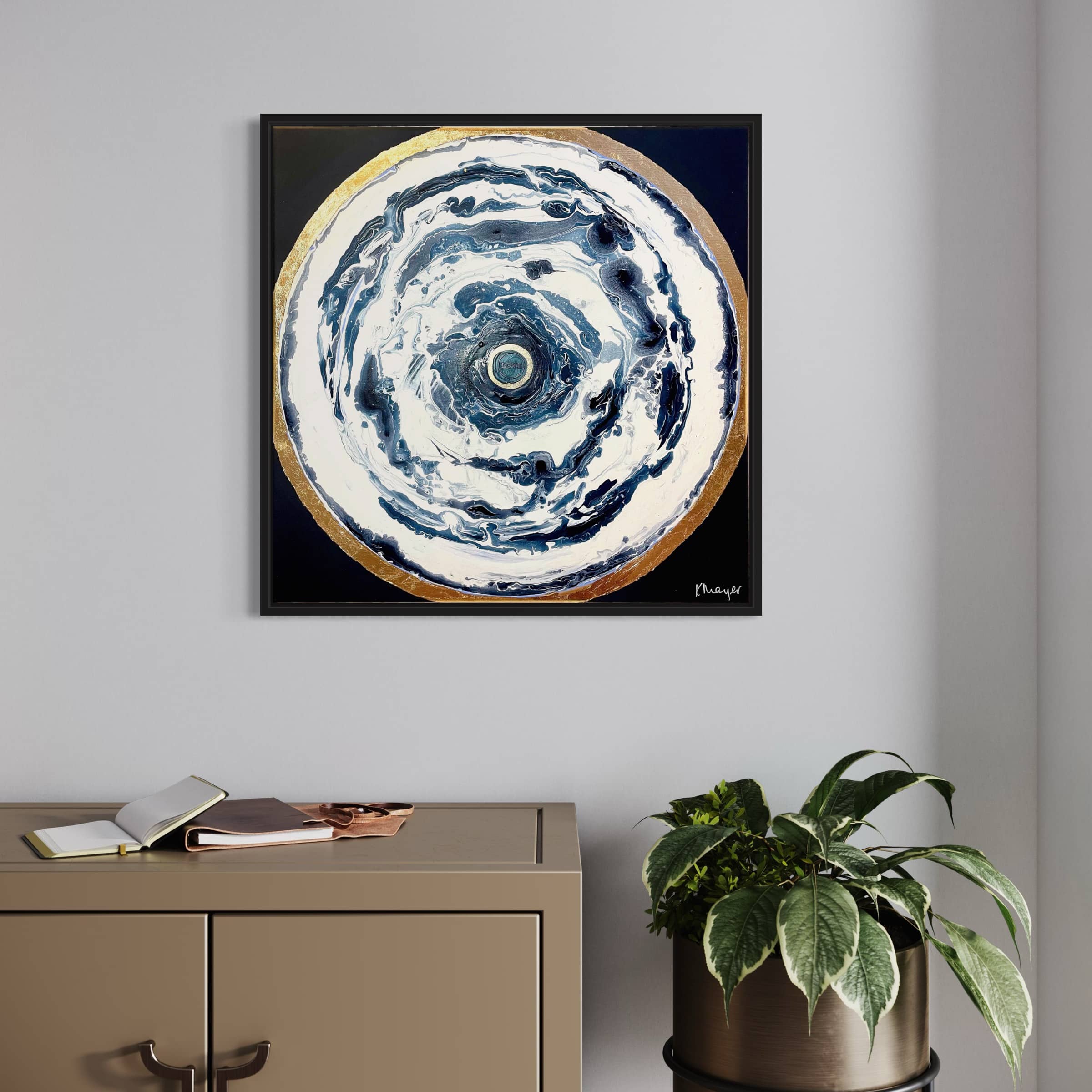 Hanging above a beige cabinet is a framed circular artwork called "Oceans of Love" by the artist Kate Mayer, accompanied by an open book and a potted plant.