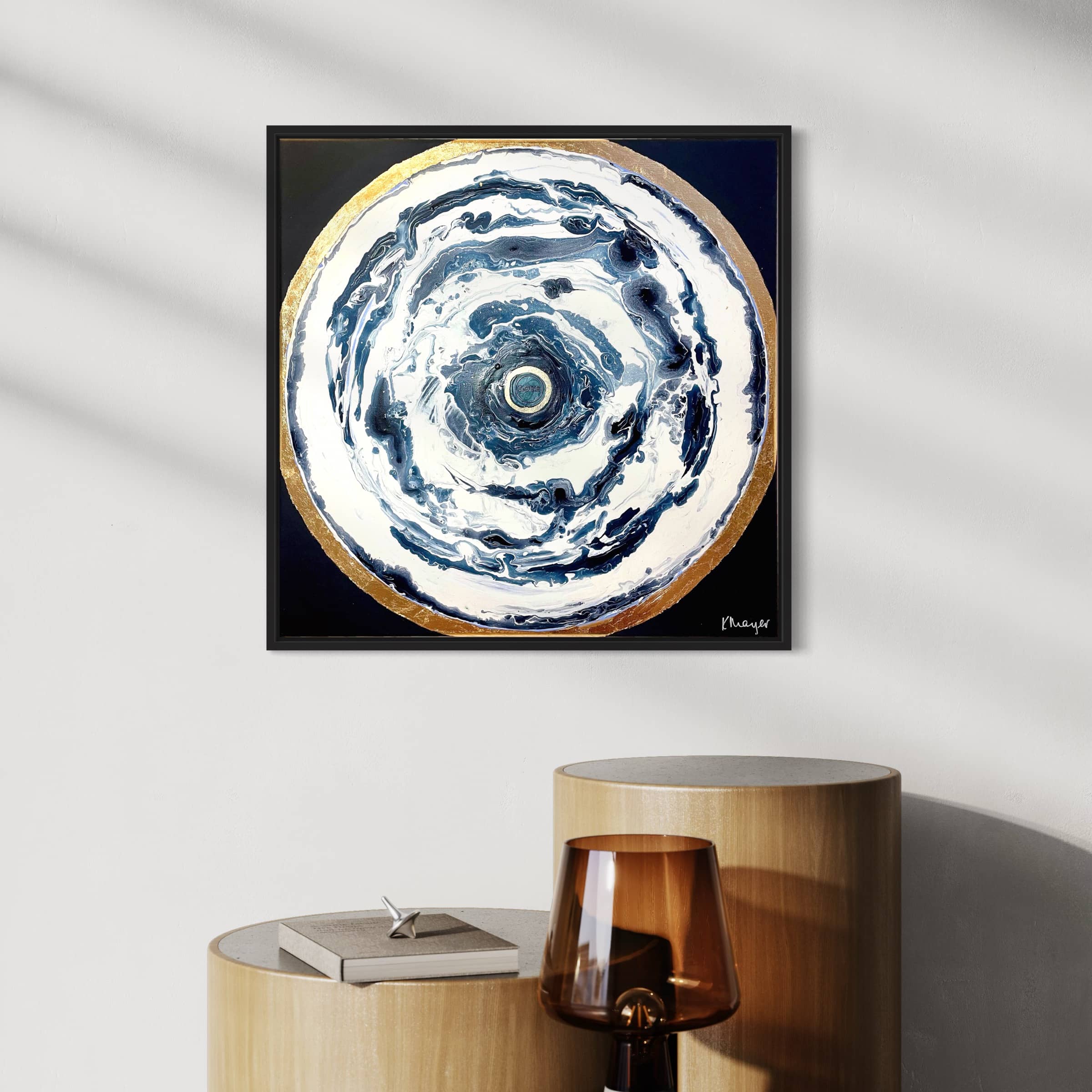 Oceans of Love," an abstract creation by Kate Mayer, enhances the wall with its circular design encased in a gold frame. Below, wooden and glass tables accentuate the artwork, one featuring a closed book, fostering a tranquil atmosphere in the room.