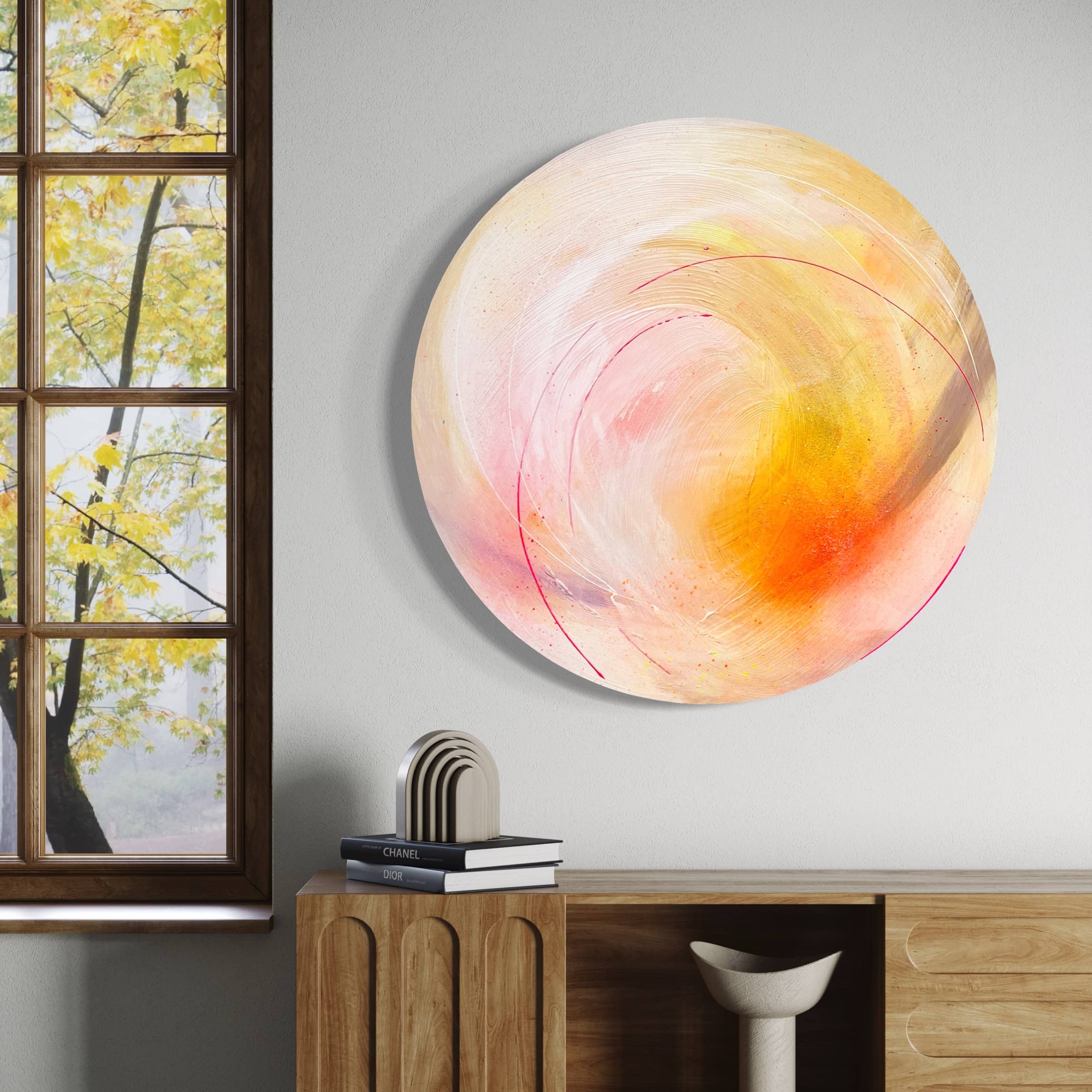 The "Happiness Is A Place Within" original painting by Kate Mayer, featuring warm colors and an abstract round design, is displayed on a wall next to a window. Beneath it, a wooden cabinet enhances the artistic ambiance of the space with neatly stacked books and decor.