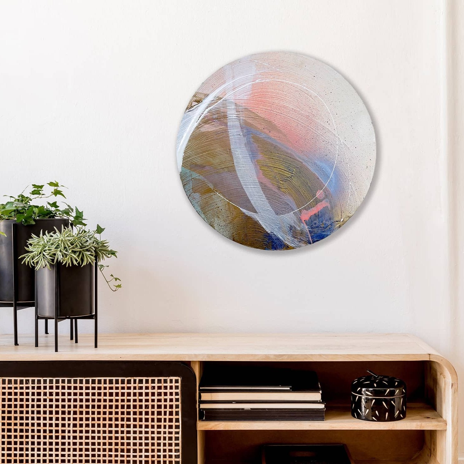 The "Just Be" painting by Kate Mayer, featuring a round design created with acrylic and spray paint in pastel hues, decorates a white wall above a wooden shelf filled with plants, books, and a small basket.