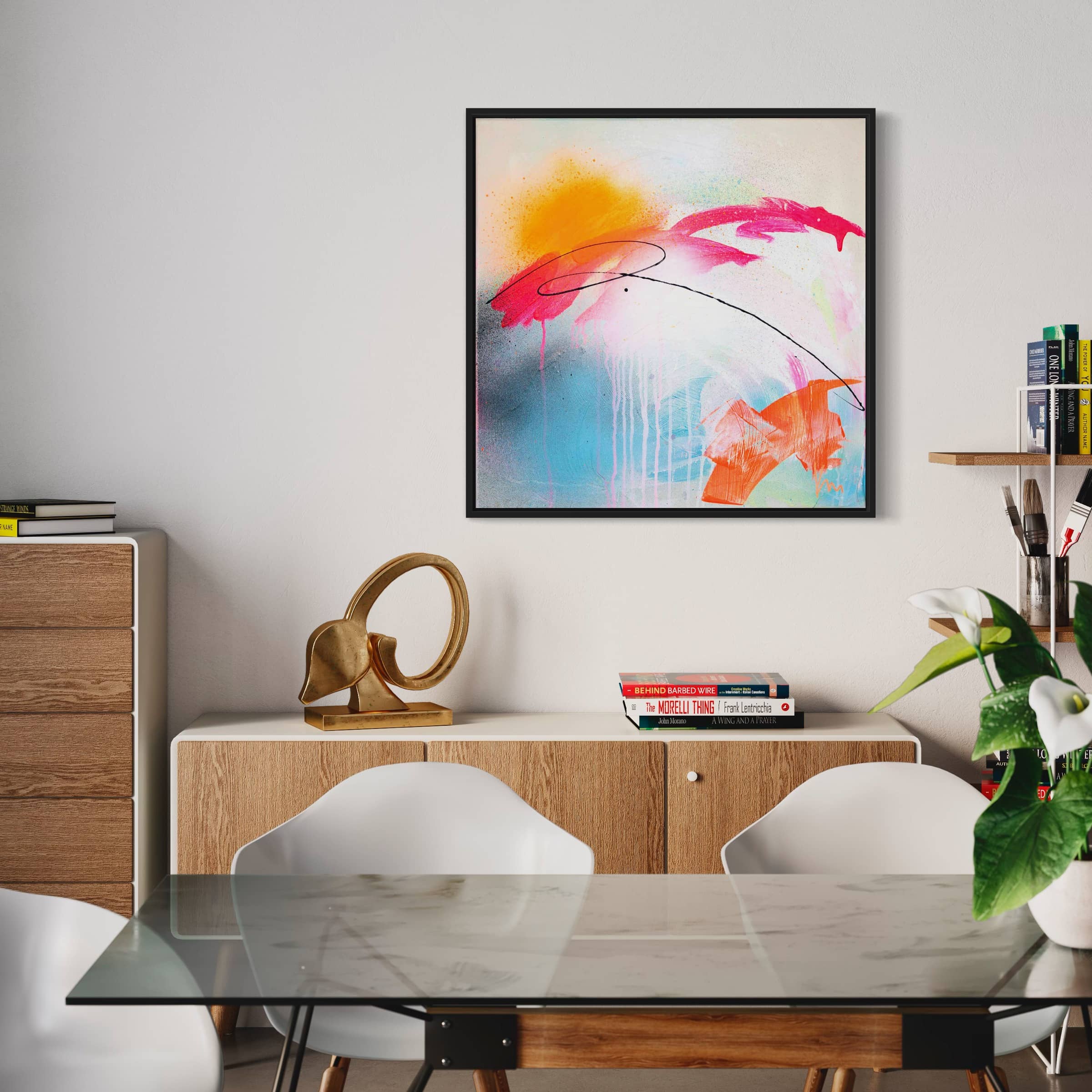 A modern dining area featuring a glass table, white chairs, and "Rise & Shine Bright," an original painting by Kate Mayer. The space is adorned with books and decorative items on elegant wooden furniture.