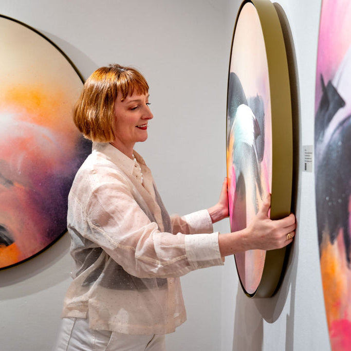 Kate Mayer adjusts "Glow Up," a vibrant round painting, on a gallery wall, wearing a sheer blouse and white pants.