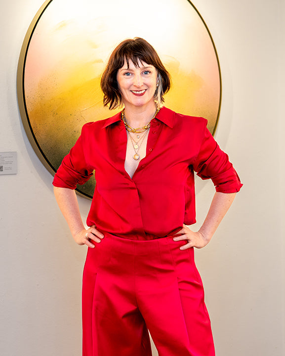 Artist Kate Mayer stands with hands on hips wearing a bright red matching two-piece outfit consisting of a long-sleeve button-up shirt and wide-leg trousers. She is posing in front of a white gallery wall decorated with three large circular abstract paintings.