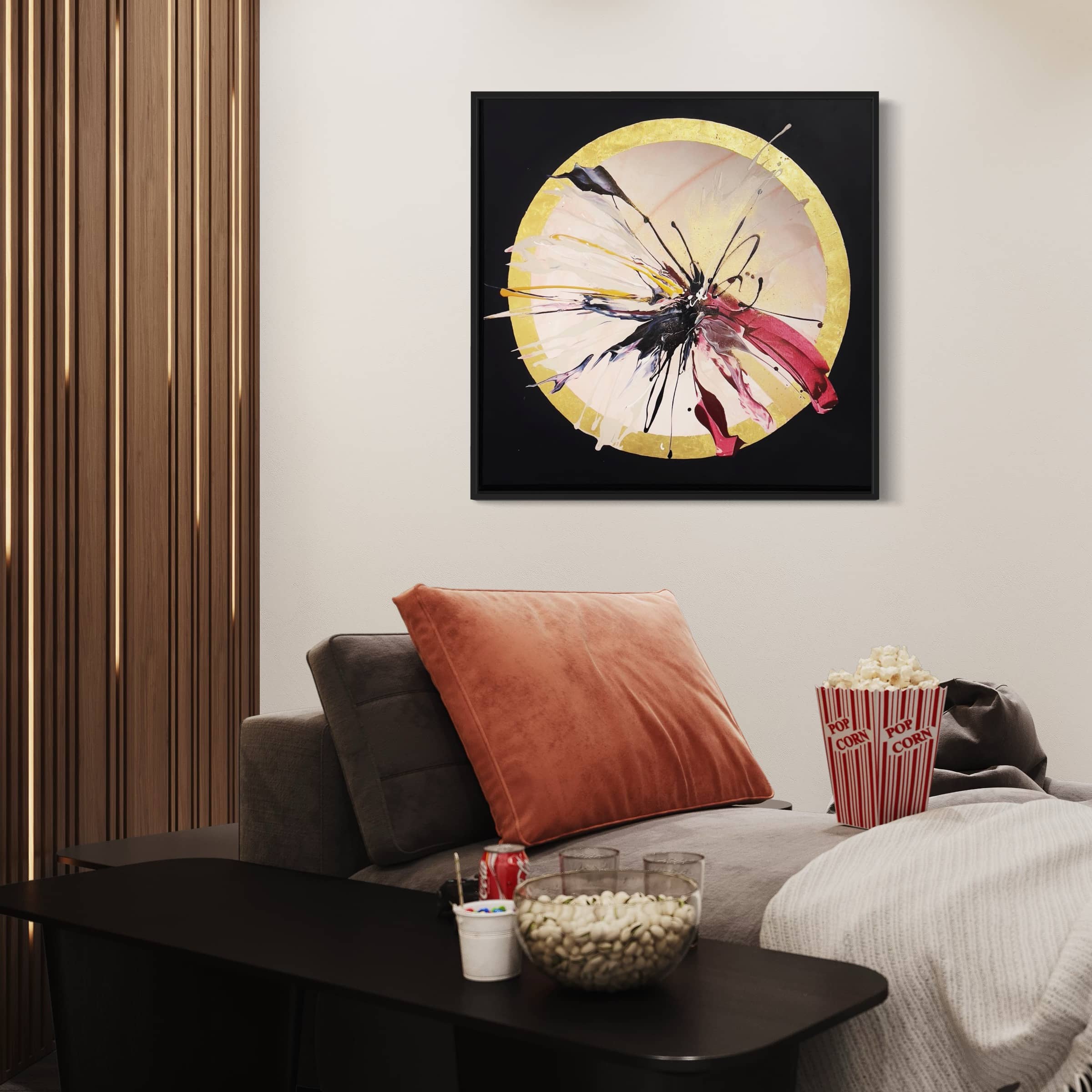 A cozy living room featuring a gray sofa, an orange pillow, and snacks is beautifully accented by "Expansions," an original painting by Kate Mayer. This ready-to-hang artwork, showcasing vibrant colors in a circular pattern with acrylic and gold leaf accents, adds a touch of elegance to the space.