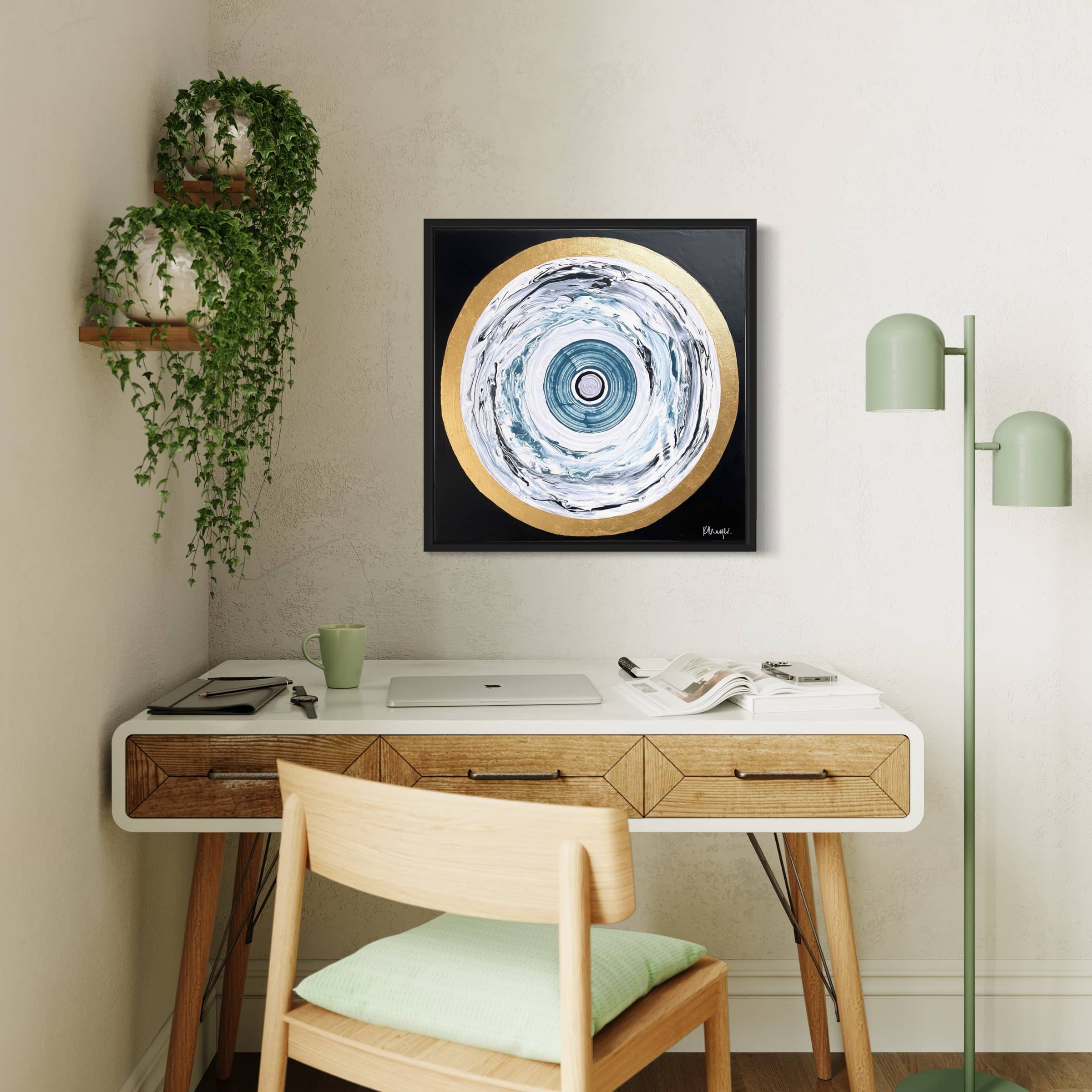 A home office with a wooden desk, white chair, laptop, green desk lamp, hanging plants, and Kate Mayer's "One Love" original abstract painting on the wall.