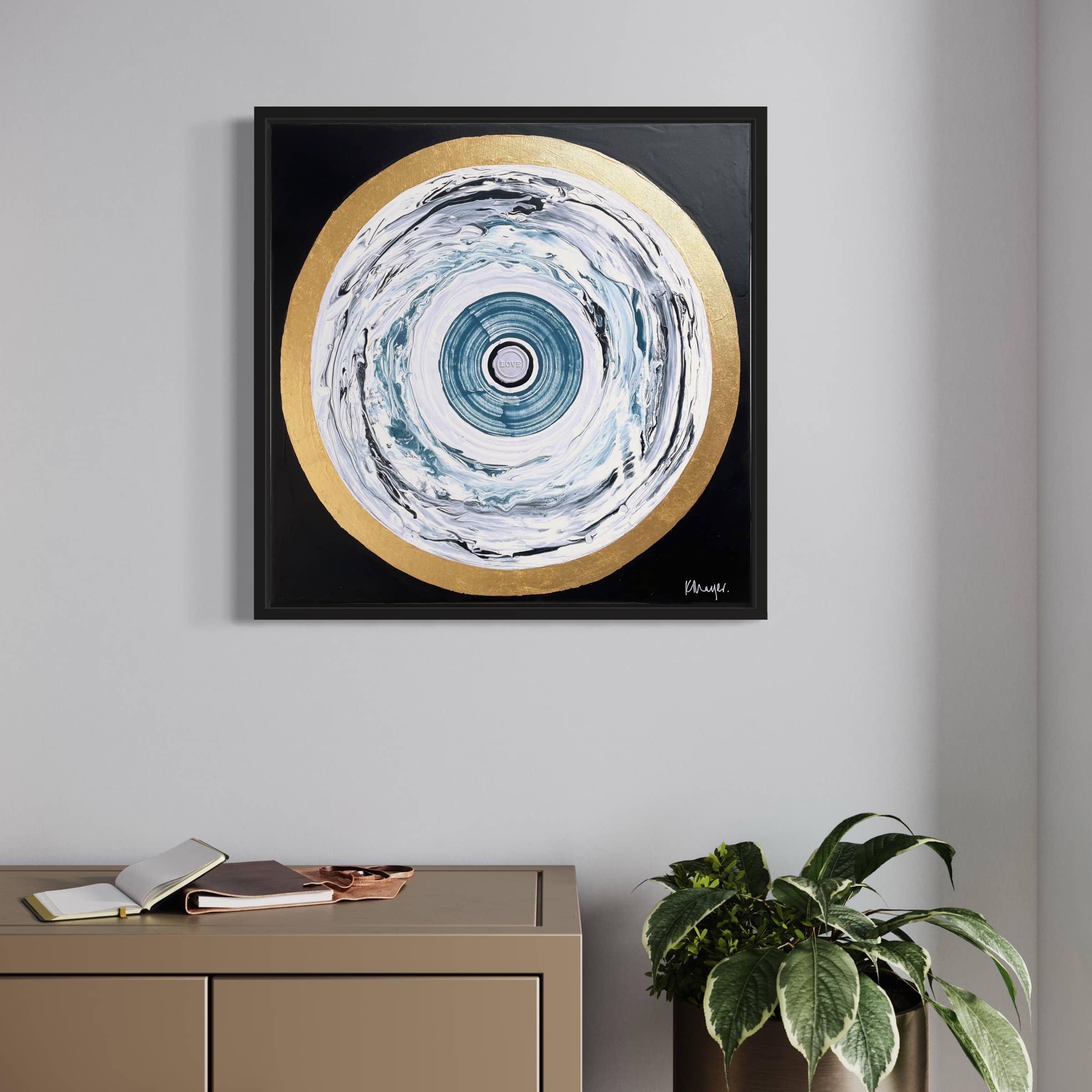 The One Love - Original Painting by Kate Mayer, featuring concentric circles in blue, white, and gold, hangs on a gray wall above a cabinet adorned with a plant and books.