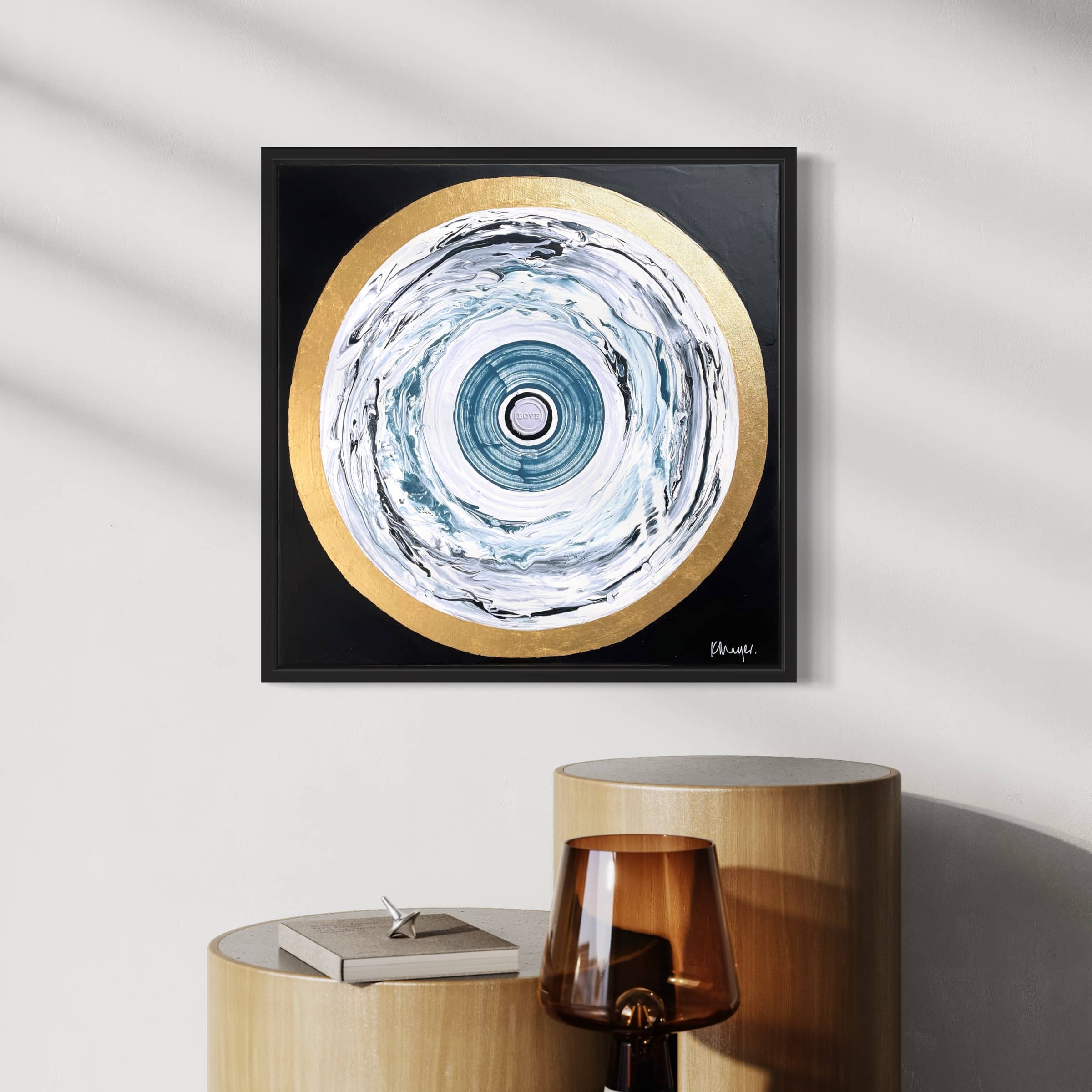 The "One Love - Original Painting" by Kate Mayer, adorned with blue, white, and gold spirals, is displayed on a white wall above beige cylindrical tables and a brown glass lamp.