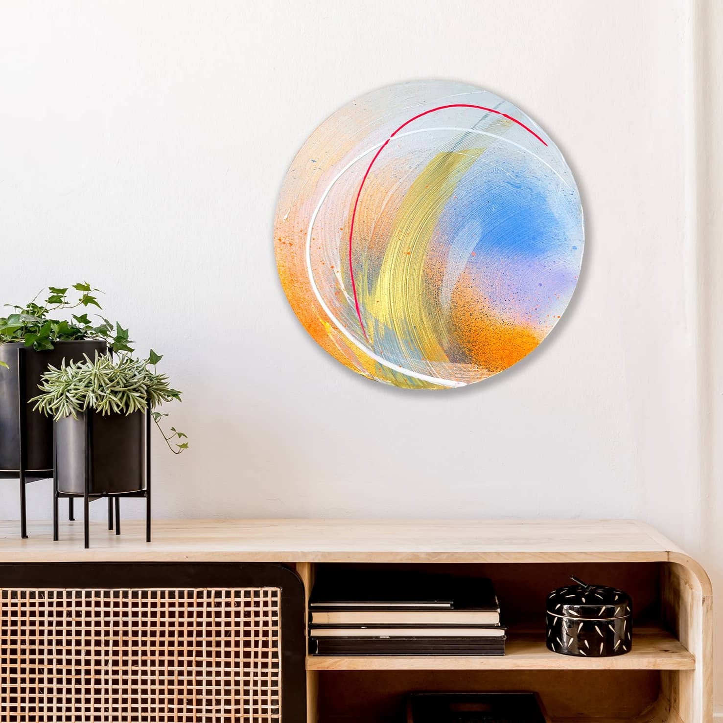 The "Be Joy" round abstract painting by Kate Mayer, featuring vibrant colors created with acrylic and spray paint, decorates the wall above a wooden cabinet adorned with plants and books.