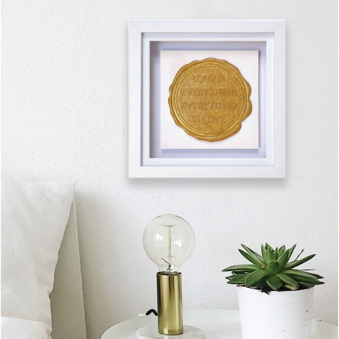 A white nightstand with a lamp and succulent plant sits beside a bed. Above the nightstand hangs "Sealed Intention for LOVE," an original piece of art by Kate Mayer, complete with a bespoke wax seal as its certificate of authentication.