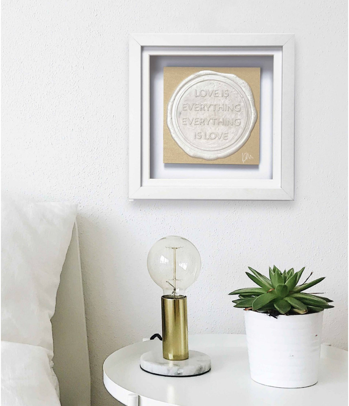 A white-framed original artwork titled "Sealed Intention for LOVE" by Kate Mayer, featuring the text "Love is Everything, Everything is Love," hangs above a round table with a lamp and potted plant. The piece comes with a certificate of authentication, ensuring its originality.