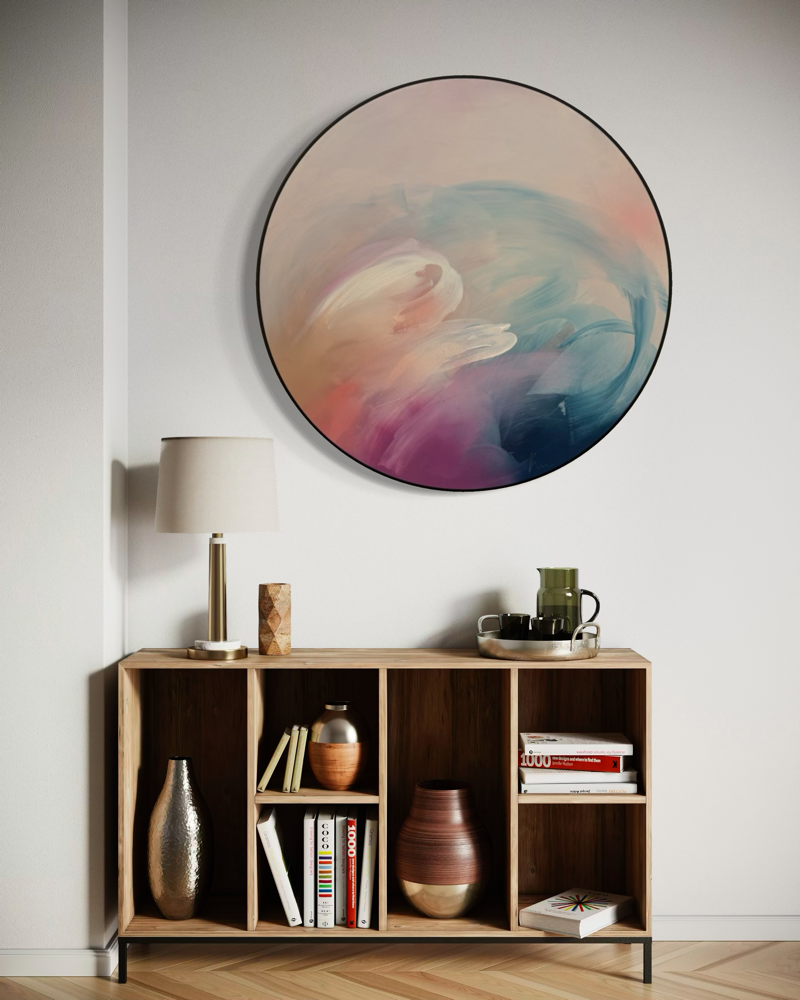 For Every Mountain“ by Kate Mayer: a round abstract painting with handmade metal frame above a cozy, book-styled shelf.
