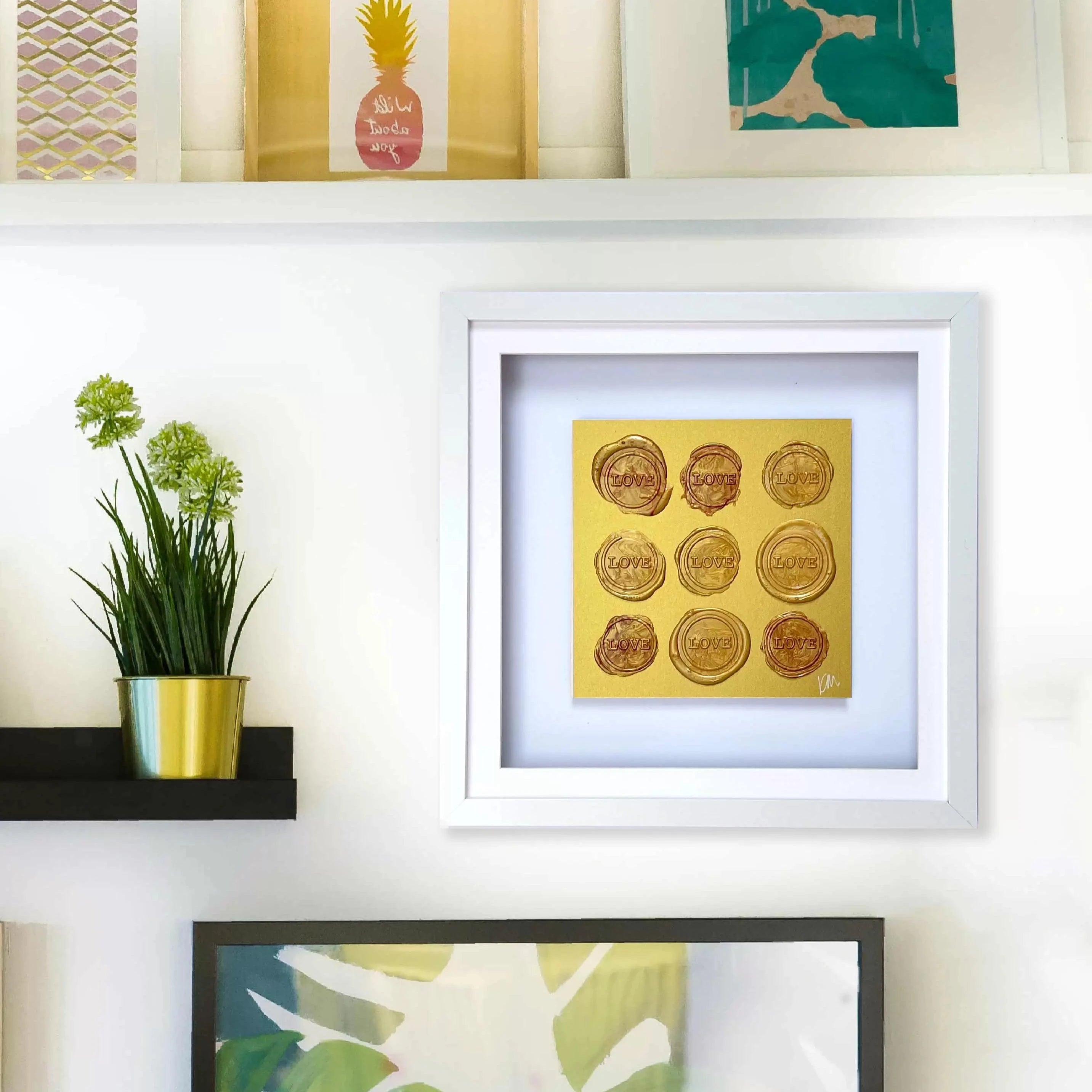 The "Sealed Intention - Love is Love" artwork, an original piece by Kate Mayer featuring gold circles on a white background and signed by the artist, is displayed on a wall shelf alongside a small potted plant.