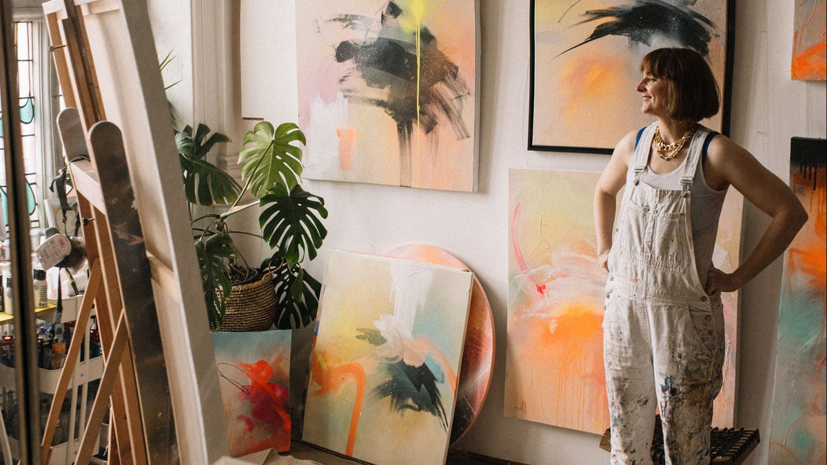 Artist Kate Mayer, n paint-splattered overalls, stands in a bright studio, surrounded by colorful abstract paintings.